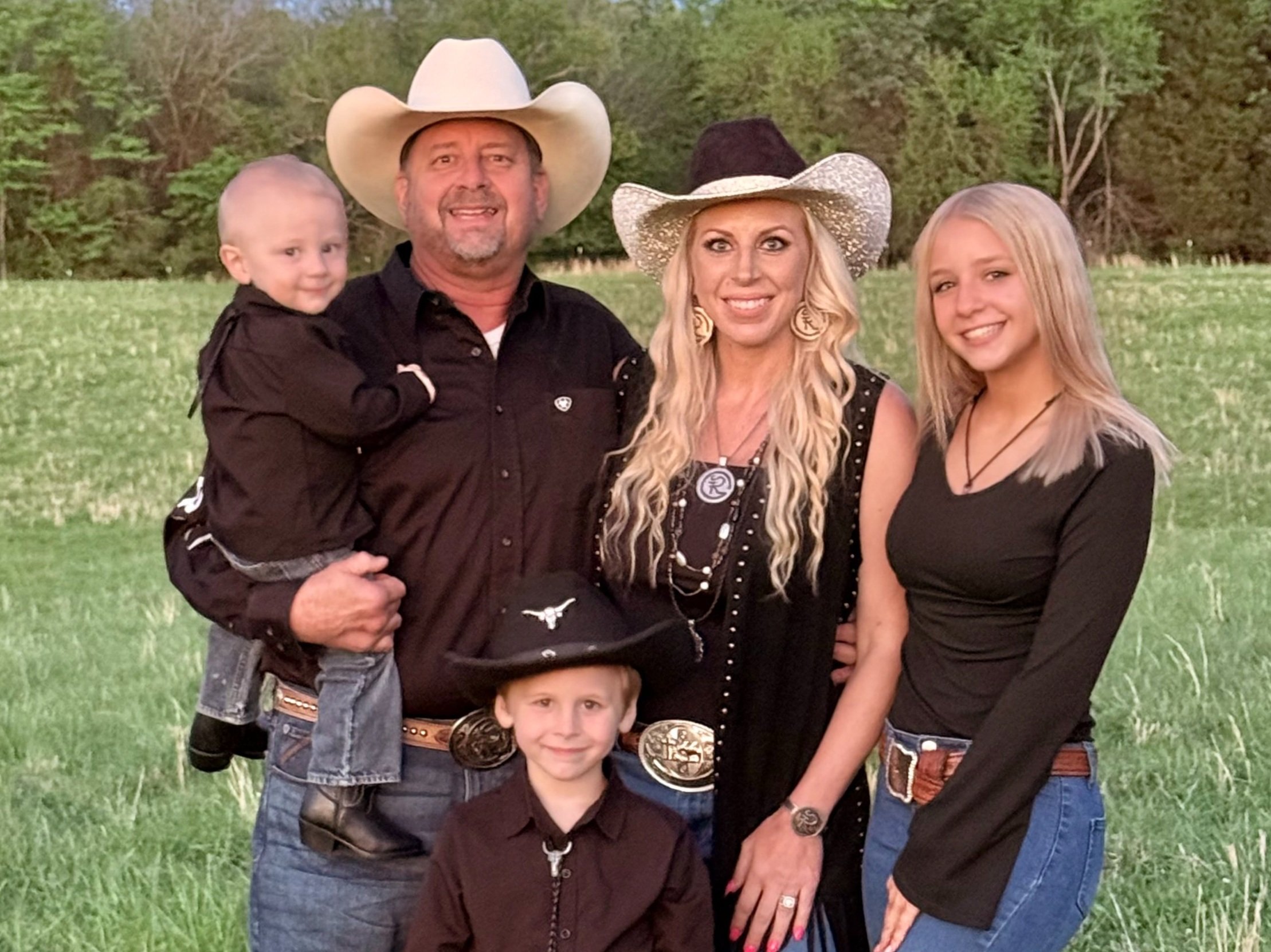 Family portrait in a rural setting, featuring a father holding a toddler, a mother, a young boy in a cowboy hat, and a teenage girl, all dressed in black outfits. The background showcases a green field and trees, capturing a warm, outdoor atmosphere.