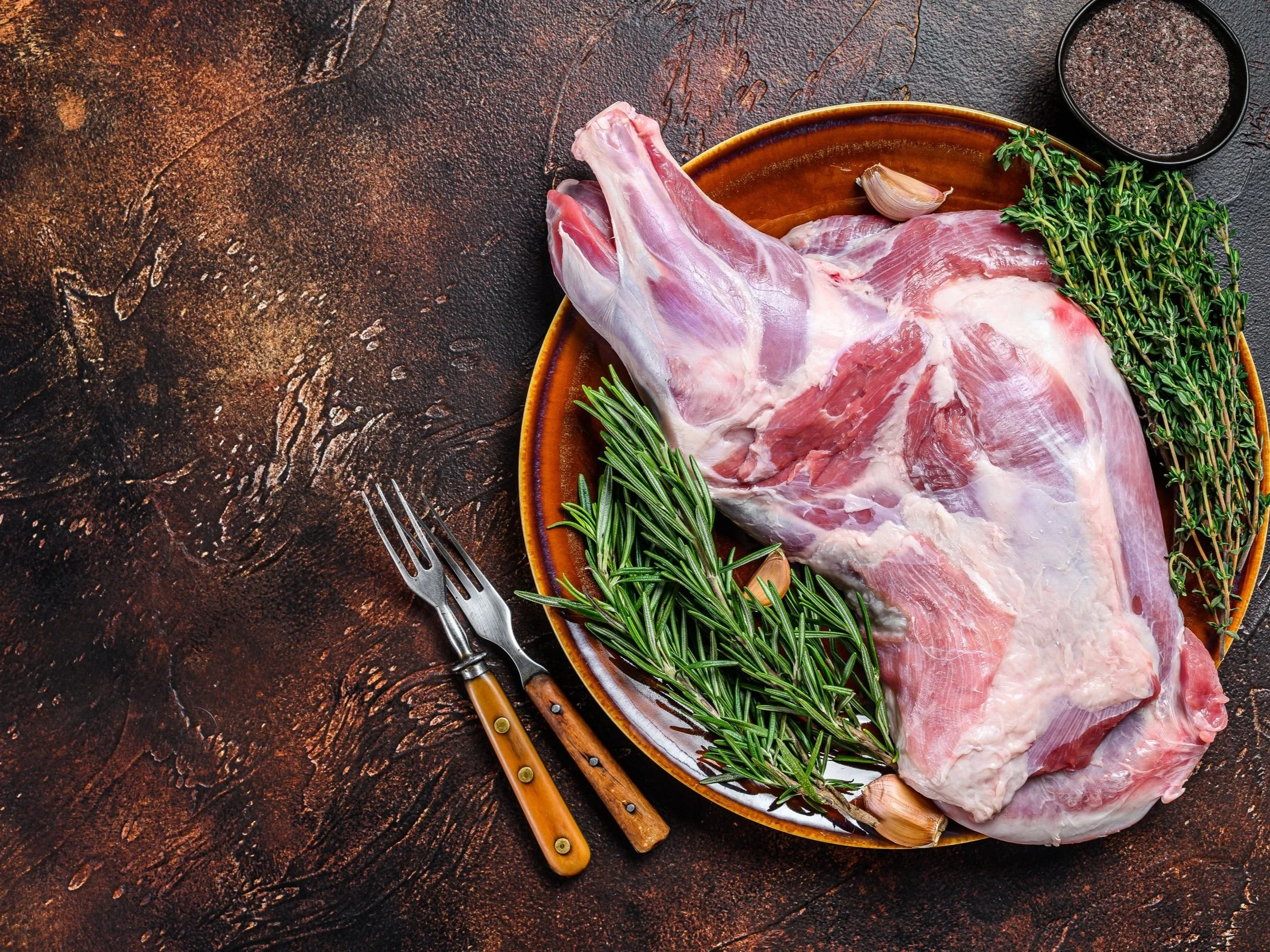 goat_shoulder Raw lamb leg displayed on a brown plate, surrounded by fresh rosemary, thyme, and garlic cloves, with two forks and a small bowl of black pepper on a textured dark background.