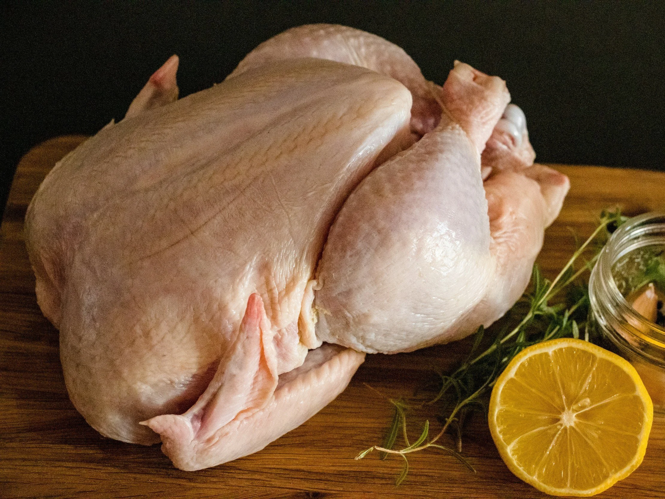 unsplash-image-9zLa37VNL38 (1) Raw whole chicken on a wooden cutting board with fresh herbs and a sliced lemon, ideal for cooking and meal preparation.
