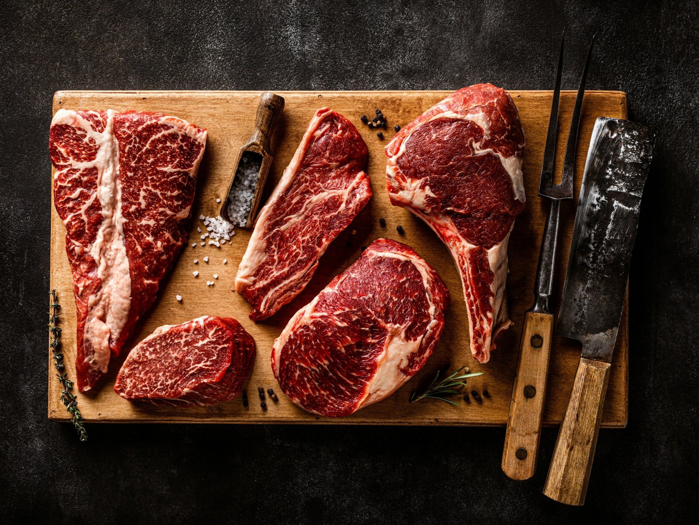 unsplash-image-cEJGctX_DgI Assorted cuts of marbled beef steak displayed on a wooden cutting board, accompanied by a small scoop of salt, herbs, and butcher tools, highlighting premium quality meat for gourmet cooking.